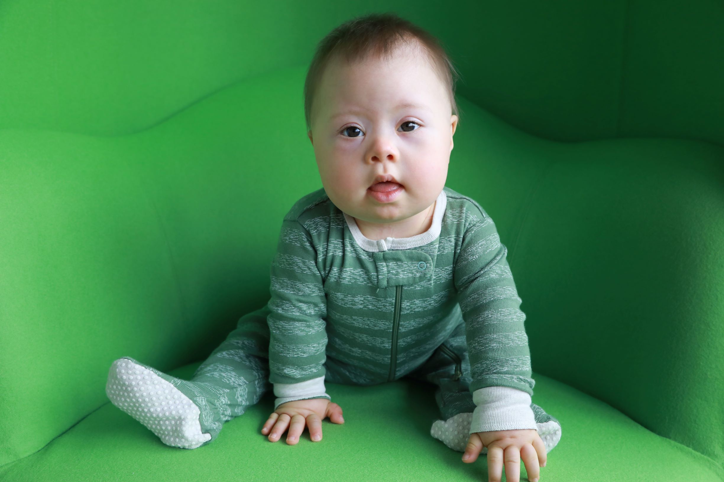 A baby with Down syndrome wearing green-striped footie pajamas sits splayed forward in a bright green chair.