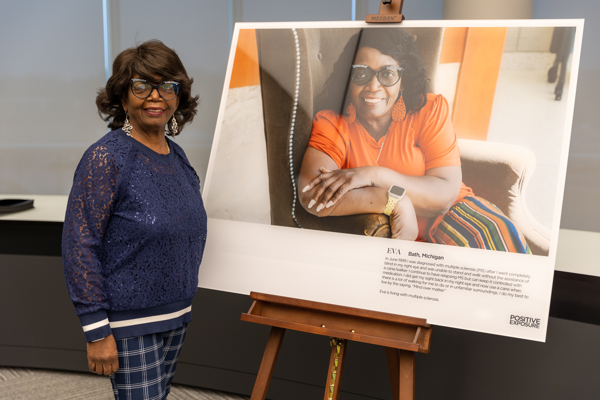 Eva, an older Black woman wearing a sparkly blue lace top and rhinestone-studded cat-eye glasses, smiles as she stands next to a large portrait of herself set on an easel. In the portrait, she's wearing the same glasses but with a bright orange top and orange earrings.